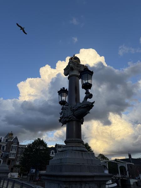 An ornate lamppost stands prominently on the Blauwbrug in Centrum, Amsterdam, silhouetted against a dramatic sky of blue and fluffy white clouds